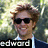 A person with light brown curly hair and dark sunglasses wears a dark suit jacket over a white collared shirt and dark tie, standing against a blurred green background with text "edward" visible.