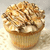 A cupcake with light brown frosting and dark brown drizzle, topped with white chunks on a textured surface.