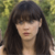 A static pixelated image of a person with long dark hair and bangs, wearing a light-colored top against a blurred green background.