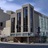 Pixelated static image of a modern multi-story building with vertical dark windows against a light blue sky background.
