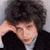 A pixelated close-up of a person with dark curly hair wearing a black jacket over a light shirt, hand raised near the head.