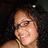 A person with curly dark hair and glasses smiles in a of four , wearing a yellow strap top against a dark background