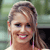 Pixelated close-up of a person with light brown hair tied back, wearing a white top with pink straps, smiling with visible teeth against a blurred green and gray background.