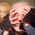Pixelated image of a person with light skin and short hair wearing a dark jacket with red trim, resting their head on a dark surface.