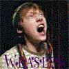 A person with light brown hair in a white shirt with purple text, mouth open wide against dark background.