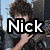 A pixelated image of a person with curly brown hair wearing dark clothing, positioned against an urban background with buildings and blue sky.