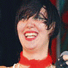A person with short dark hair and red lipstick smiles widely, wearing a white top with black-and-white striped ruffles and a red choker.