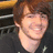 A pixelated image of a person with brown hair and a black shirt on a pink and gray background with red text.
