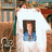 A person holds a white T-shirt with a printed portrait of another individual against a bookshelf background.