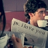 A person with curly dark hair holds a newspaper with 'Ps. I love you' printed on it, while another person holds a cup in a dimly lit indoor setting.