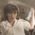 A pixelated figure with dark hair wears a light-colored shirt and holds a light blue object against a muted brown background.