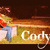 Pixelated image of a person with brown hair in yellow top and blue pants holding an orange object, next to white text reading 'Cody' on golden background.