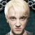 Pixelated close-up of a person with short light-colored hair, neutral expression, and visible facial features against a dark background.
