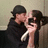 Two figures in dark clothing kiss while one holds a silver microphone against a light wall background.