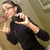 Pixelated image of a person with dark hair wearing a black sleeveless top, making a hand gesture with fingers extended and thumb up against a muted brown background.