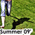 Pixelated image of a person in light blue shorts and white socks standing on green grass with a black dog lying nearby, text "SUMMER 09" at bottom.