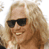 A person with long blonde hair and dark sunglasses smiles, wearing a white shirt with a black-and-white winged cross logo reading "TOWN DOG".