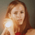 A pixelated close-up of a person with long brown hair wearing a red top, holding a bright glowing object near their shoulder against a muted gray background.