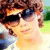 Pixelated image of a person with brown hair wearing dark sunglasses, light-colored shirt, and blue jacket, standing outdoors with green and blue background.