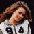 Static pixelated image of a person with long brown hair wearing a white shirt with black text, set against dark background.