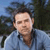 Pixelated image of a person in a light blue shirt with a dark collar, set against a blurred background of blue sky and dark green foliage.