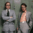 Two men in gray suits stand against a muted green background; one has arms crossed, the other's jacket is open revealing his chest.
