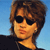 Pixelated portrait of a person with brown hair wearing dark sunglasses and a black top against a blue background.