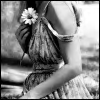 Black-and-white image of a person with long hair tied back by a flower, wearing a sleeveless dress with a patterned skirt and a wide belt.