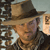 A pixelated static image shows a person wearing a brown wide-brimmed hat and dark clothing, with a light-colored background featuring partial text elements.