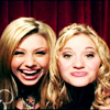 Two blonde women with wavy hair pose side-by-side against a red curtain background, one smiling broadly with teeth visible, the other with a closed-mouth smile.