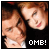 Two pixelated figures with light brown hair and fair skin appear in close proximity, one slightly behind the other, against a dark background with white text reading "OME!" at bottom right.