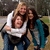 Three people embrace in a close group hug; one wears white, another dark clothing with teal accents, and the third has long brown hair.