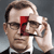 Pixelated image of a person with light brown hair wearing dark glasses and a dark suit, holding a white object near their face against a gray background.