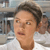Pixelated close-up of a person with short dark hair wearing a white shirt, set against a blurred background with indistinct shapes and colors.