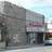 Pixelated static image of a building facade with a red sign, dark entrance, and light-colored ground under a pale sky.