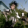 A person with dark tousled hair in a white shirt and suspenders moves through green foliage against a light blue building background.