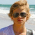 A person with light brown hair and dark sunglasses stands against a beach background with waves and a clear sky.