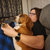 A pixelated image shows a person with light skin and dark hair holding a small brown and white dog against a blurred background.