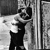 Black-and-white image of two people embracing against a textured wall, one person in a sleeveless top and dark pants, the other in a dark dress, both with arms around each other.