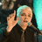 A pixelated image of a person with light hair in a dark jacket, holding a microphone, against a blue background with red text.