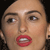Static pixelated image of a person's face with light skin tone, brown eyebrows, brown eyes, and red lips showing teeth, set against a dark background.