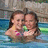 Two people in a swimming pool, one with long brown hair wearing a pink bikini top, the other with short brown hair, both facing forward with neutral expressions.