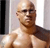 Pixelated image of a bald man with dark skin tone, wearing dark sunglasses, standing in front of a light-colored wall with vertical dark and light stripes.