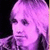Pixelated image of a person with light purple hair and dark eyes, wearing a dark patterned garment against a black background.