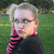 Pixelated image of a person with light-colored hair tied back, wearing dark glasses and a black top with pink stripes on the sleeves, standing against a green grassy background.