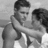 Two grayscale figures in close physical proximity, one with short dark hair and a beard wearing a sleeveless top, the other with shorter dark hair partially visible.
