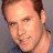 A pixelated image of a person with short brown hair and light skin, wearing a dark blue shirt against a gray background.