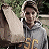 A person in a gray hoodie holds up a bagel with both hands, standing outdoors against greenery and a light-colored structure.