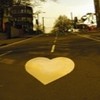 A heart-shaped icon with a yellow gradient sits on a blurred urban street background with buildings and trees.