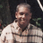 Static pixelated image of a person with short light hair wearing a light-colored collared shirt against a dark background with green foliage.